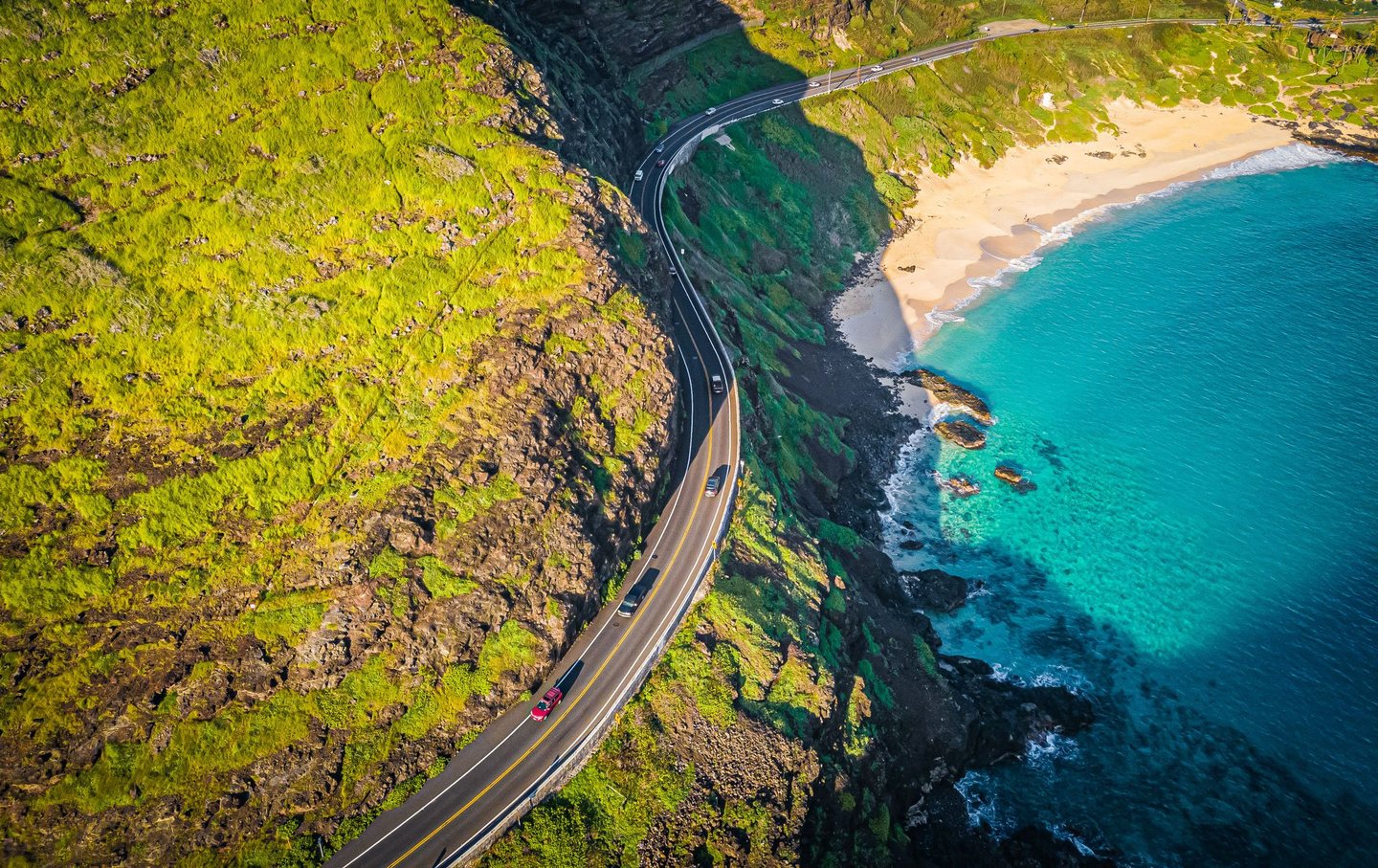 An aerial view shows cars following a winding road between steep green cliffs and turquoise ocean water beside a sandy beach.