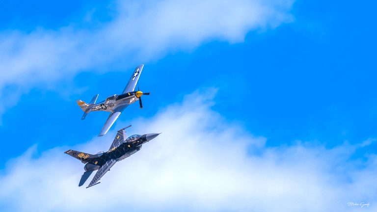 A silver propeller fighter and a dark patterned jet bank together against a bright blue sky with white clouds.