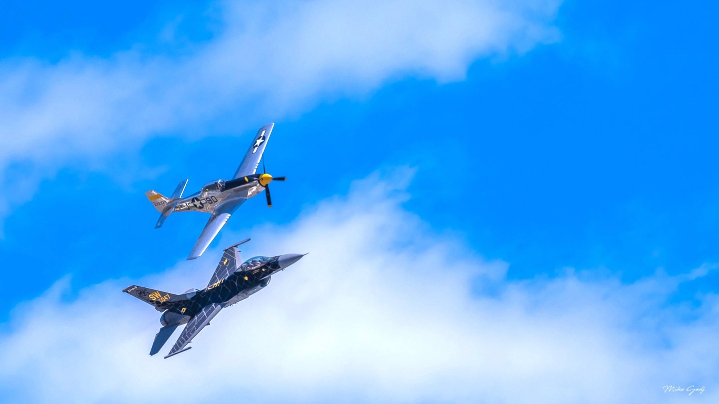 A silver propeller fighter and a dark patterned jet bank together against a bright blue sky with white clouds.