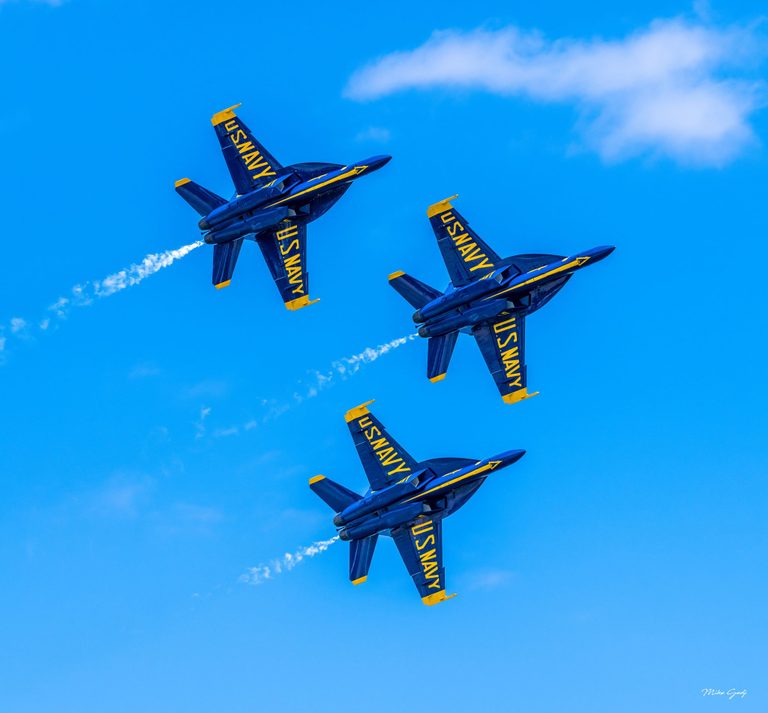 Three blue and yellow U.S. Navy Blue Angels jets bank in formation against a bright blue sky, trailing thin streams of white smoke.