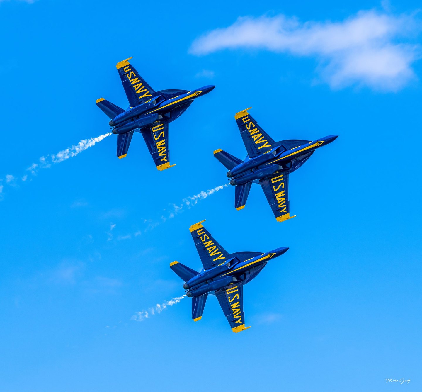 Three blue and yellow U.S. Navy Blue Angels jets bank in formation against a bright blue sky, trailing thin streams of white smoke.