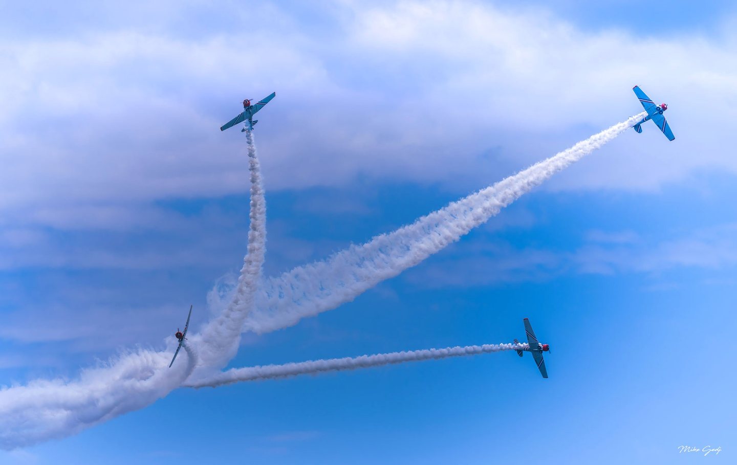 Four propeller aircraft bank in different directions, leaving diverging white smoke trails across a blue sky scattered with clouds.