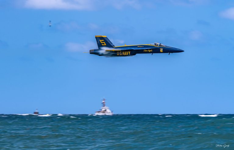 A blue and yellow U.S. Navy Blue Angels jet marked number 5 flies above choppy ocean water, with boats along the horizon.
