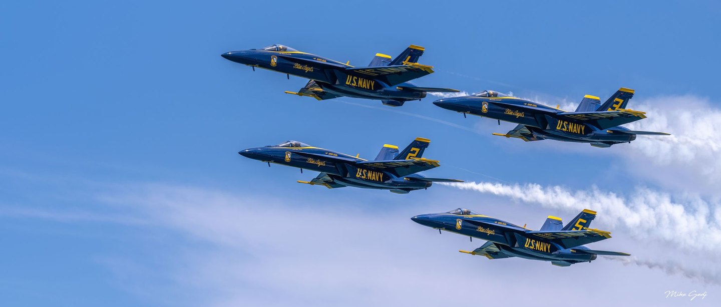 Four blue and yellow U.S. Navy Blue Angels jets fly leftward in formation against a blue sky, with white smoke trailing behind them.