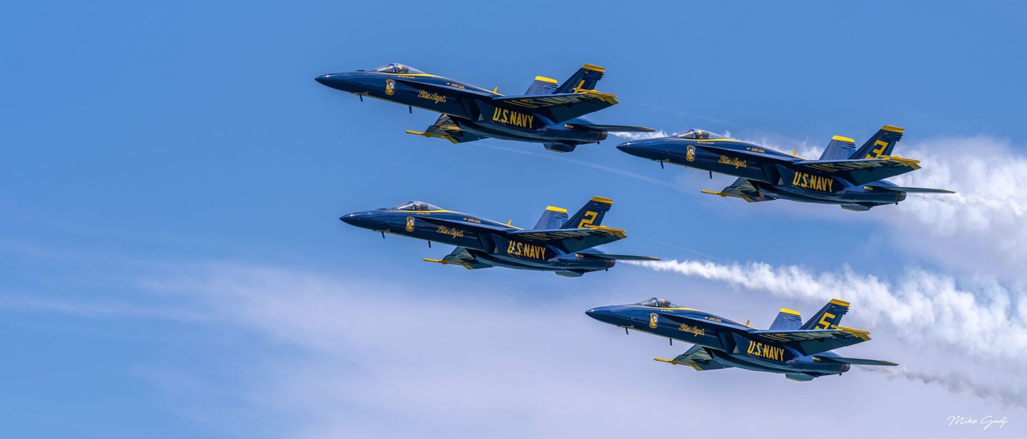 Four blue and yellow U.S. Navy Blue Angels jets fly leftward in formation against a blue sky, with white smoke trailing behind them.