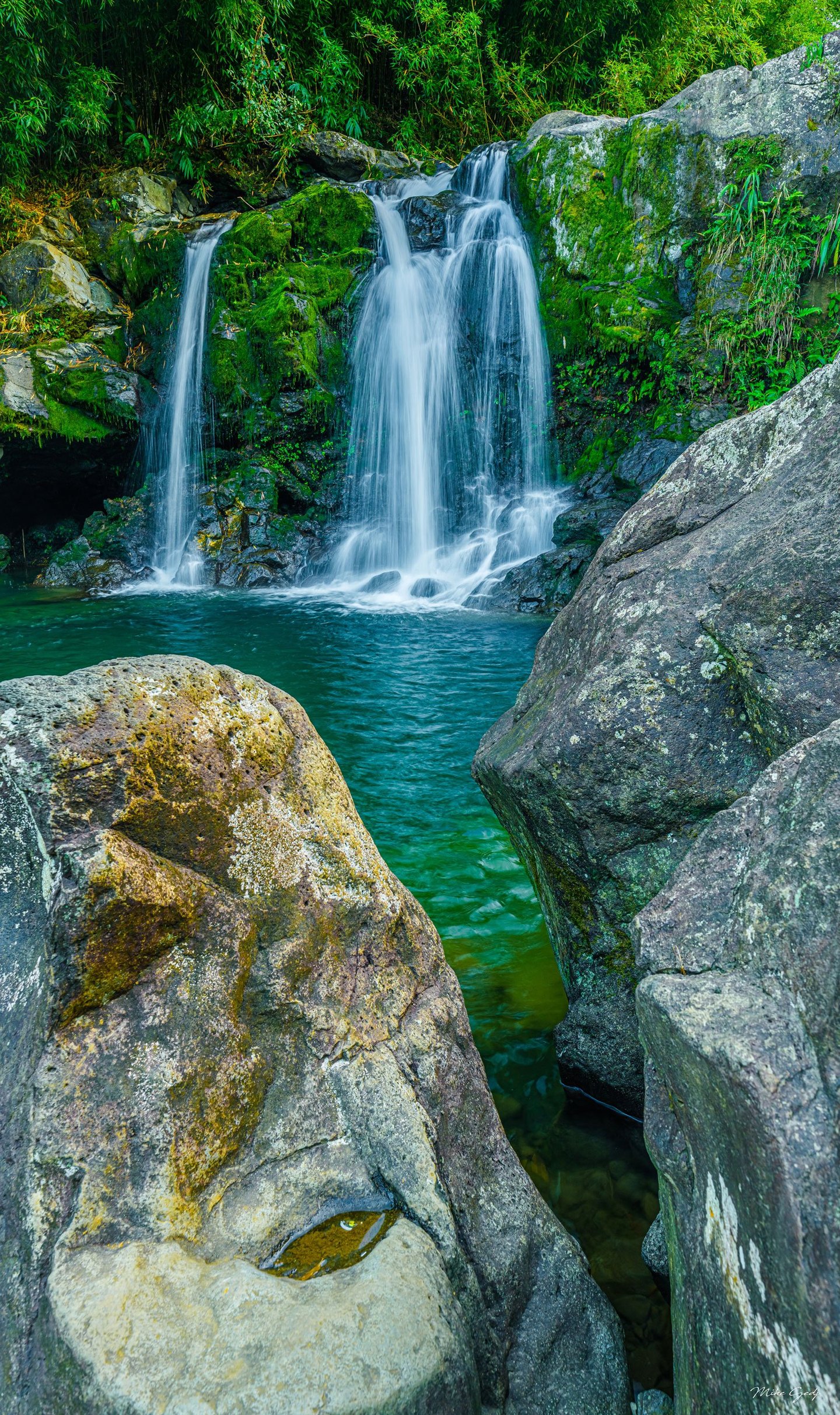 A waterfall cascades over moss-covered rocks into a turquoise pool framed by large gray boulders and dense green foliage.