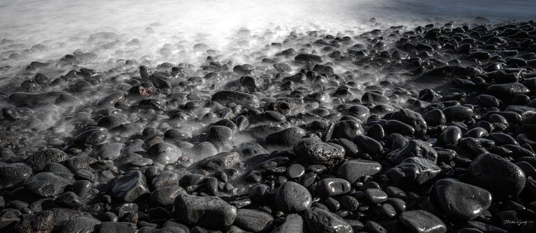 Silky white surf washes between dark, wet stones on a rocky shoreline, with bright reflections fading into deep foreground shadows.
