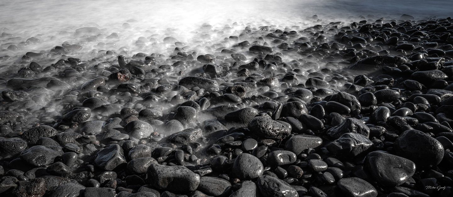 Silky white surf washes between dark, wet stones on a rocky shoreline, with bright reflections fading into deep foreground shadows.