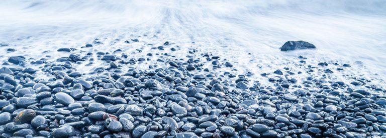 Pale blue surf washes over a shore of smooth, dark pebbles, with a larger rock emerging from the water near the upper right.