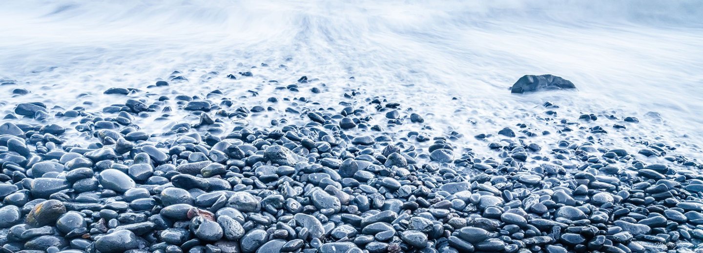 Pale blue surf washes over a shore of smooth, dark pebbles, with a larger rock emerging from the water near the upper right.