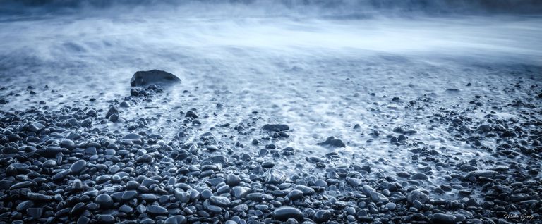 Blue-toned surf washes over a pebble beach, with softly blurred water surrounding dark rounded stones and a larger rock left of center.