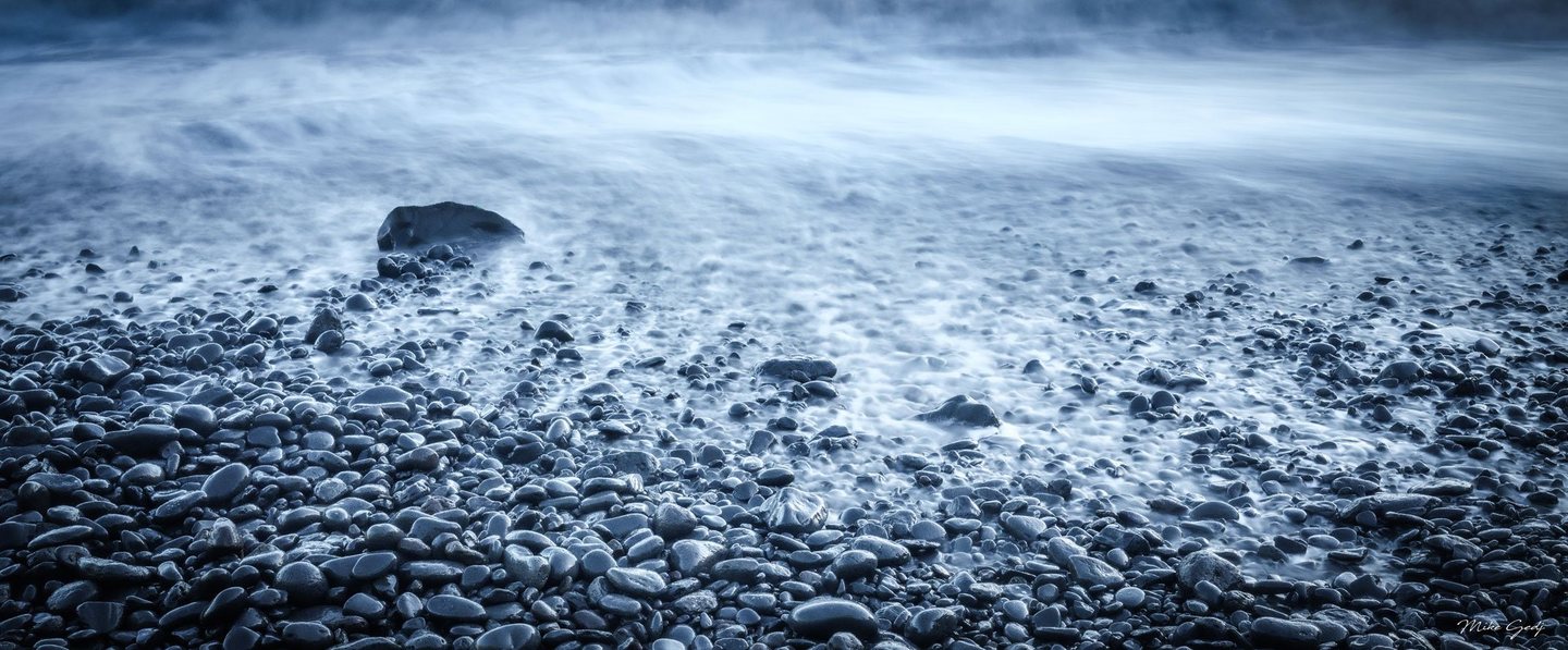 Blue-toned surf washes over a pebble beach, with softly blurred water surrounding dark rounded stones and a larger rock left of center.
