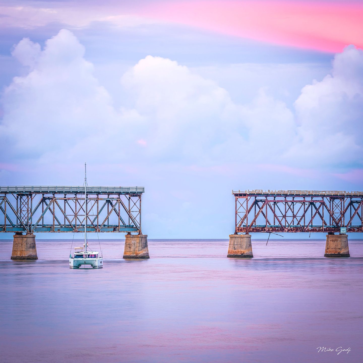A white catamaran floats on calm pink and lavender water in front of two separated rusted bridge spans beneath towering pale clouds.