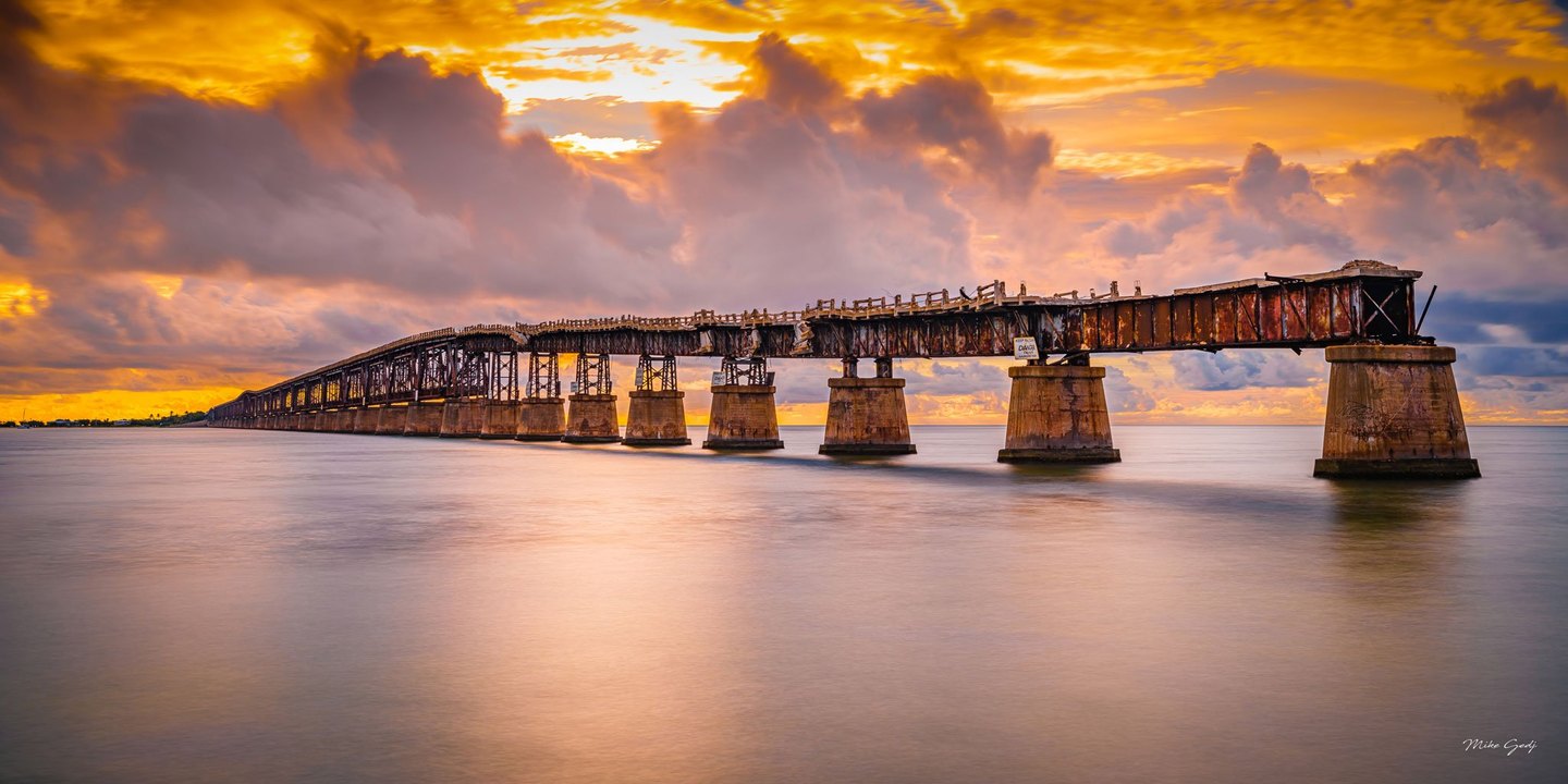 A rusted, partially ruined steel bridge stretches across calm water on concrete piers beneath vivid golden clouds.