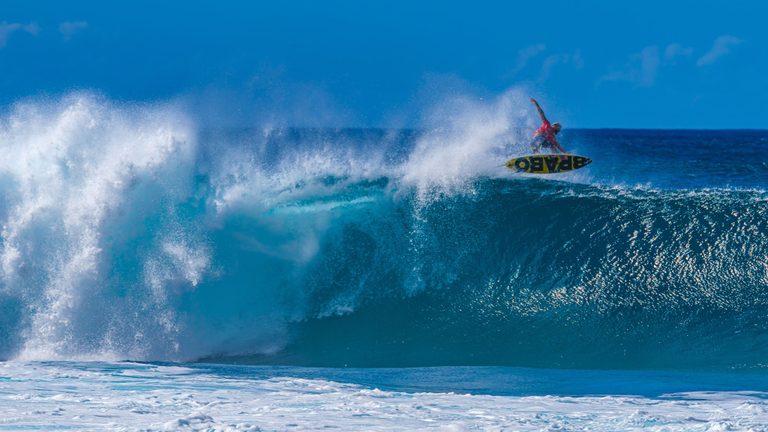 A surfer in a red shirt lifts a yellow-and-black board above the crest of a large turquoise wave amid white spray.