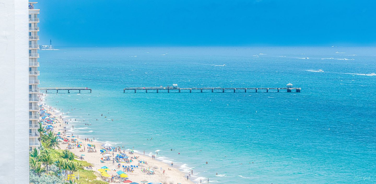 An elevated view shows a busy sandy beach with colorful umbrellas and palm trees beside a white high-rise, with a long pier and boat wakes across turquoise water.
