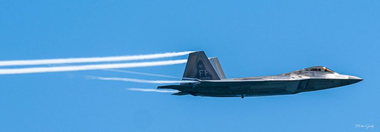 A gray F-22 Raptor fighter jet flies toward the right against a clear blue sky, with white vapor trails streaming behind it.