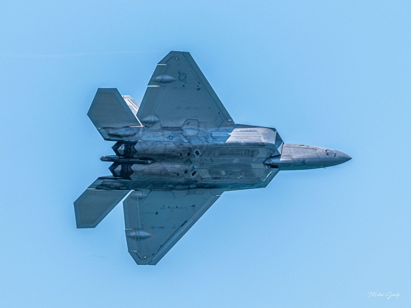 A gray F-22 Raptor fighter jet banks against a clear blue sky, revealing its underside, angular wings, and twin exhausts.