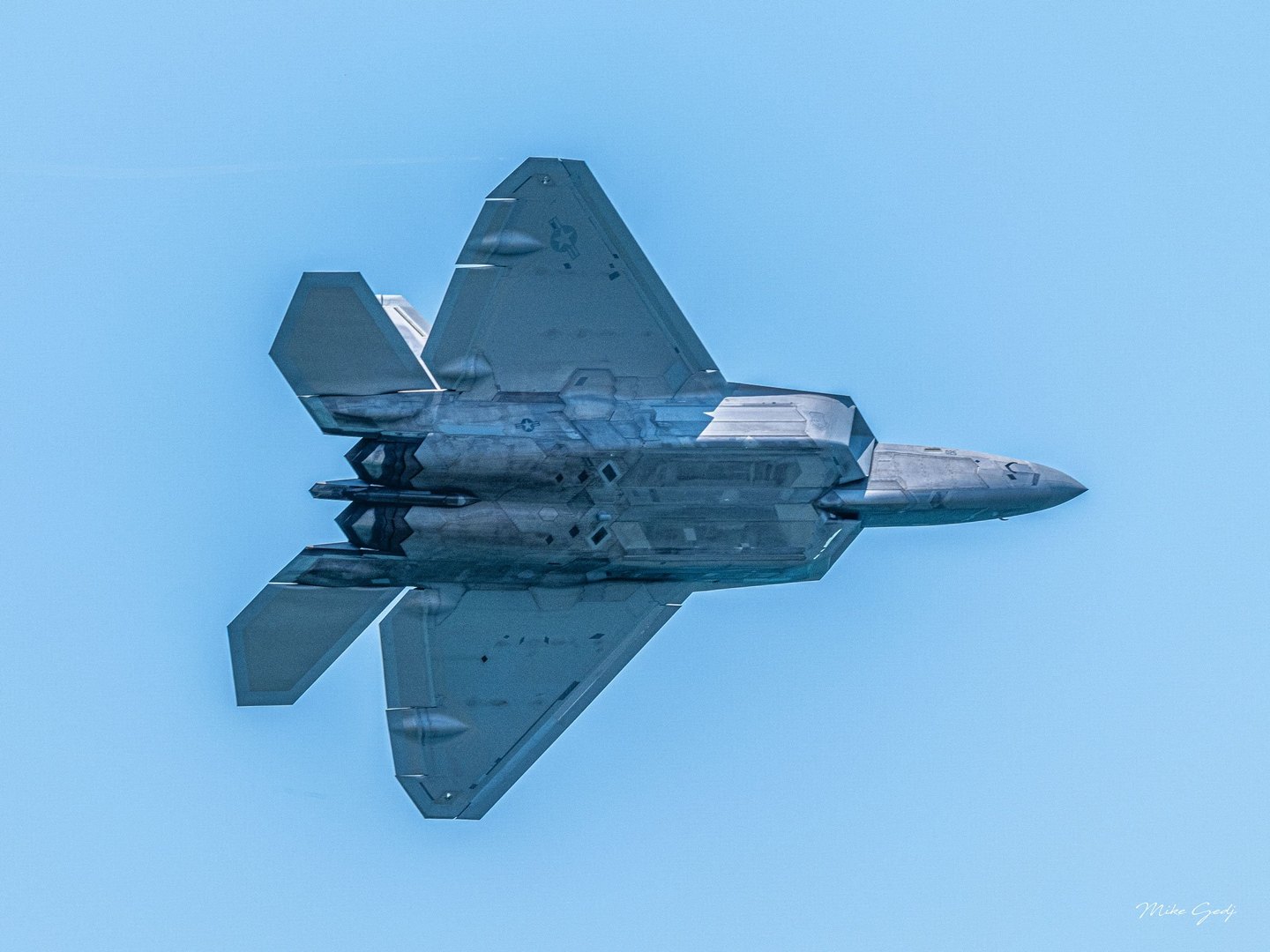 A gray F-22 Raptor fighter jet banks against a clear blue sky, revealing its underside, angular wings, and twin exhausts.