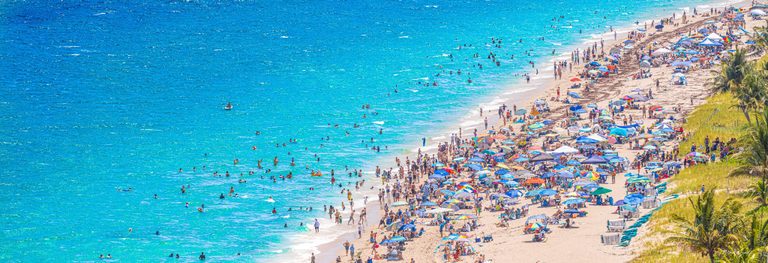 An elevated view shows swimmers in turquoise water beside a crowded sandy beach lined with colorful umbrellas, lounge chairs, dune grass, and palms.