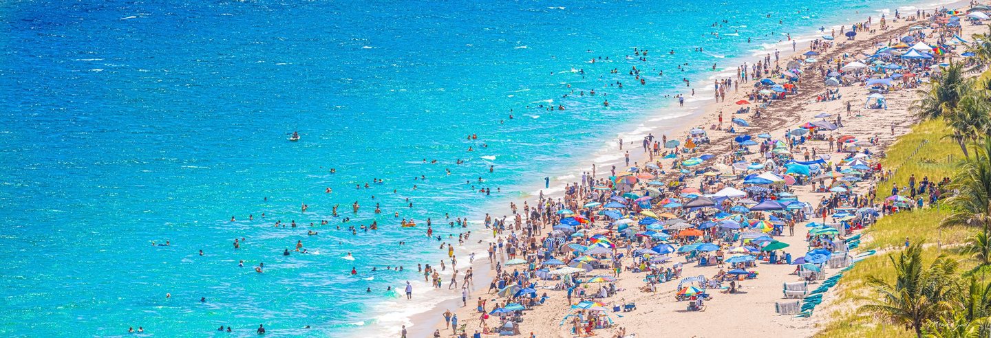 An elevated view shows swimmers in turquoise water beside a crowded sandy beach lined with colorful umbrellas, lounge chairs, dune grass, and palms.