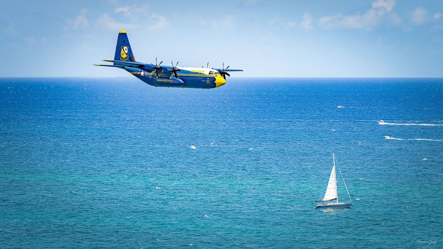 A blue and yellow Blue Angels transport aircraft flies above a turquoise sea, with a white sailboat below and small motorboats leaving wakes to the right.