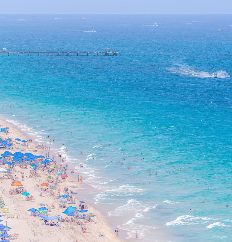 An elevated view shows a crowded sandy beach with colorful umbrellas, swimmers in turquoise water, a long pier, and boats scattered across the blue sea.