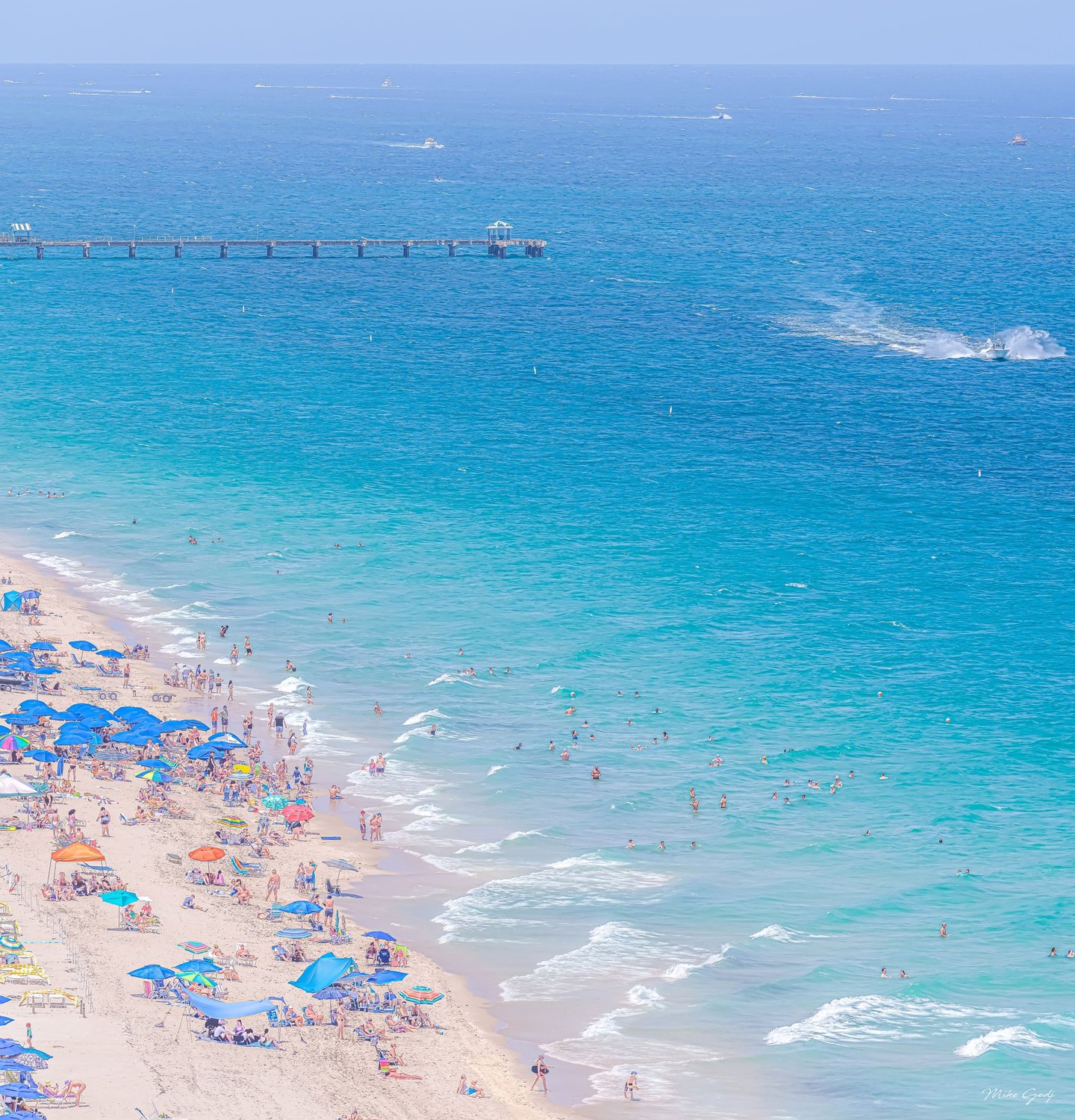 An elevated view shows a crowded sandy beach with colorful umbrellas, swimmers in turquoise water, a long pier, and boats scattered across the blue sea.