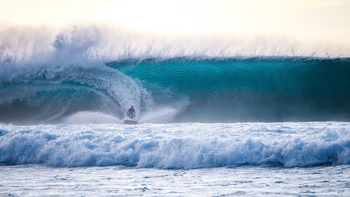 A rider on a jet ski crosses foaming surf beneath a towering turquoise wave breaking into white spray.