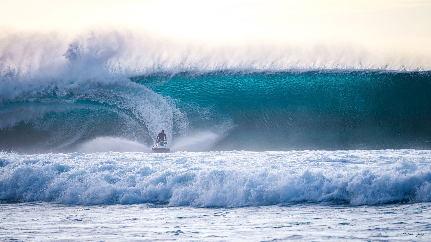 A rider on a jet ski crosses foaming surf beneath a towering turquoise wave breaking into white spray.