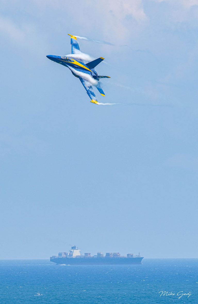 A blue and yellow jet banks steeply with white vapor curling over its wings above a distant container ship on the blue sea.