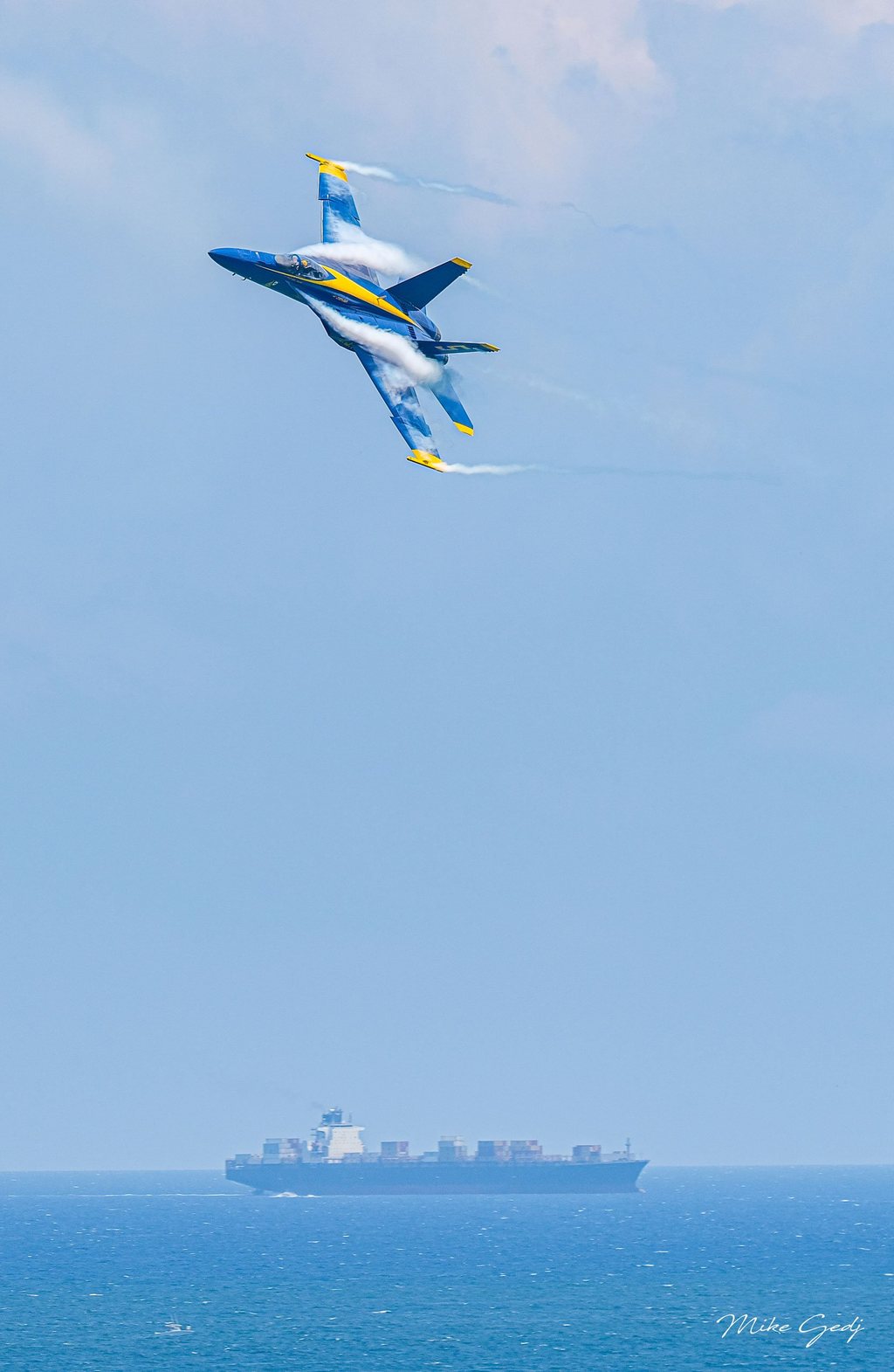 A blue and yellow jet banks steeply with white vapor curling over its wings above a distant container ship on the blue sea.
