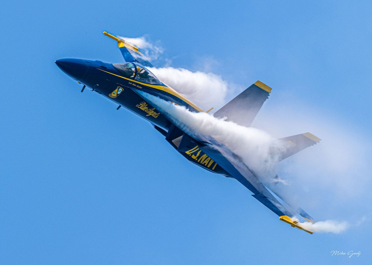 A blue and yellow U.S. Navy Blue Angels jet banks against a clear blue sky with white condensation streaming across its wings.