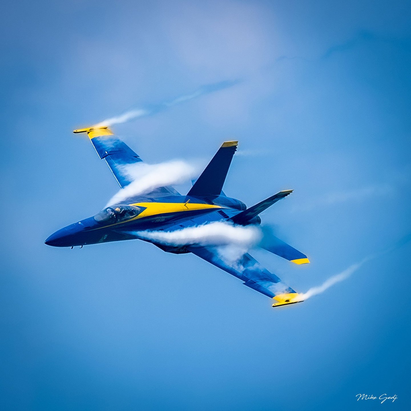 A blue and yellow fighter jet banks against a blue sky, with white condensation streaming over its wings and trailing from its wingtips.
