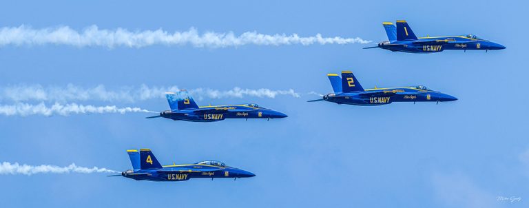 Four blue and yellow U.S. Navy Blue Angels jets fly toward the right in a staggered formation, trailing white smoke across a blue sky.