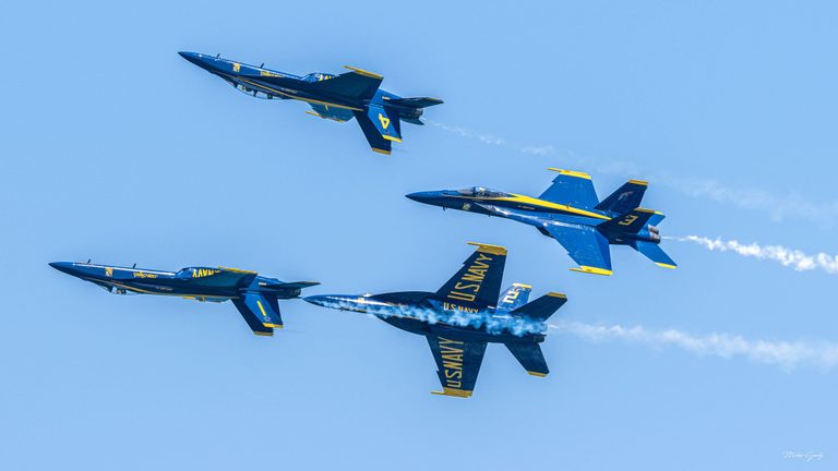 Four blue and yellow U.S. Navy Blue Angels jets fly at different bank angles against a clear blue sky, with white smoke trailing behind them.