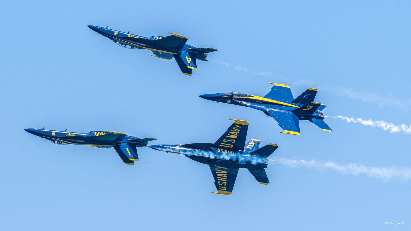 Four blue and yellow U.S. Navy Blue Angels jets fly at different bank angles against a clear blue sky, with white smoke trailing behind them.