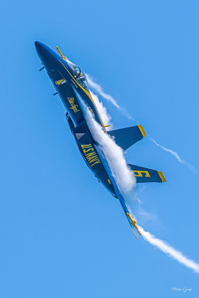 A blue and yellow U.S. Navy Blue Angels jet banks steeply against a clear blue sky, with white vapor curling over its wings.