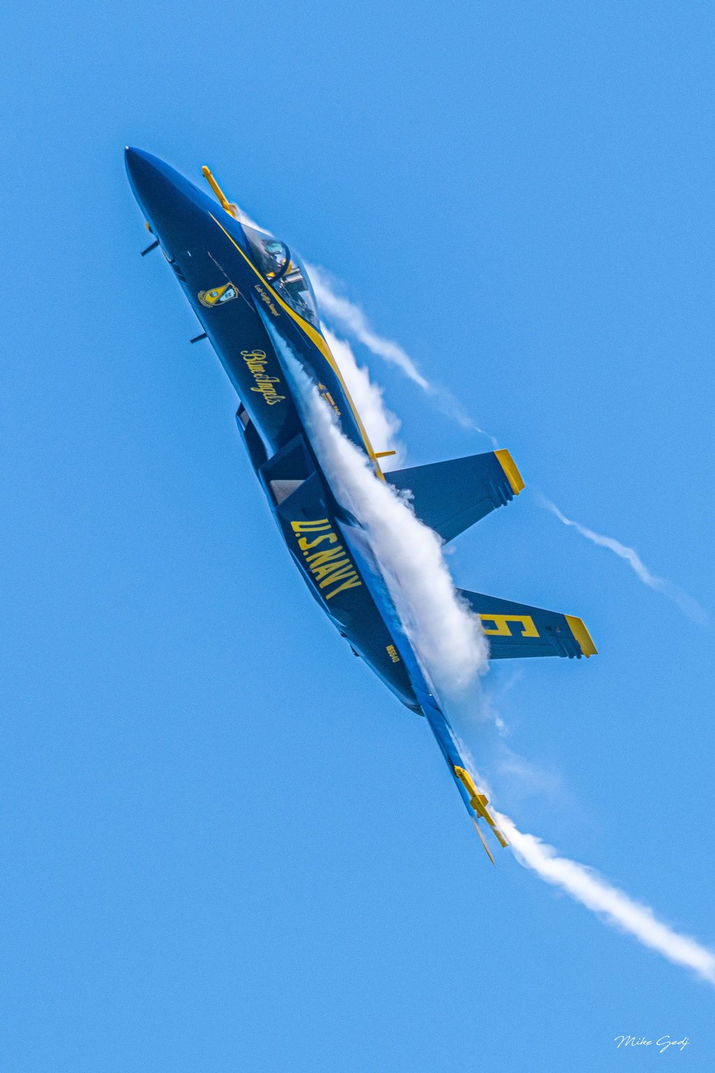 A blue and yellow U.S. Navy Blue Angels jet banks steeply against a clear blue sky, with white vapor curling over its wings.