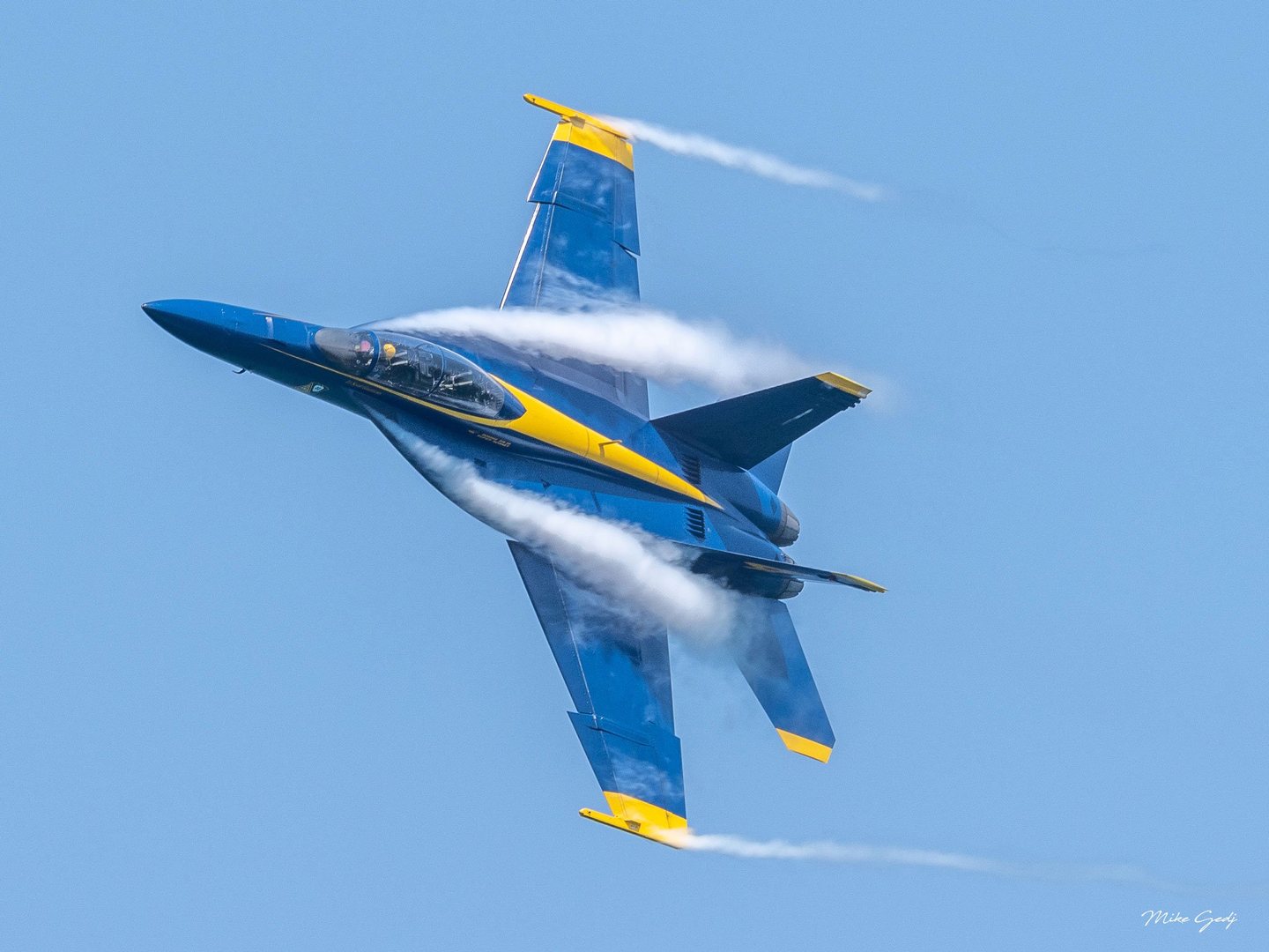 A blue and yellow Blue Angels jet banks steeply against a clear blue sky, with white condensation sweeping across its wings and trailing from its wingtips.