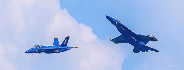 Two blue and yellow U.S. Navy Blue Angels jets fly against a pale blue sky and white clouds, one level on the left and the other banking steeply on the right.