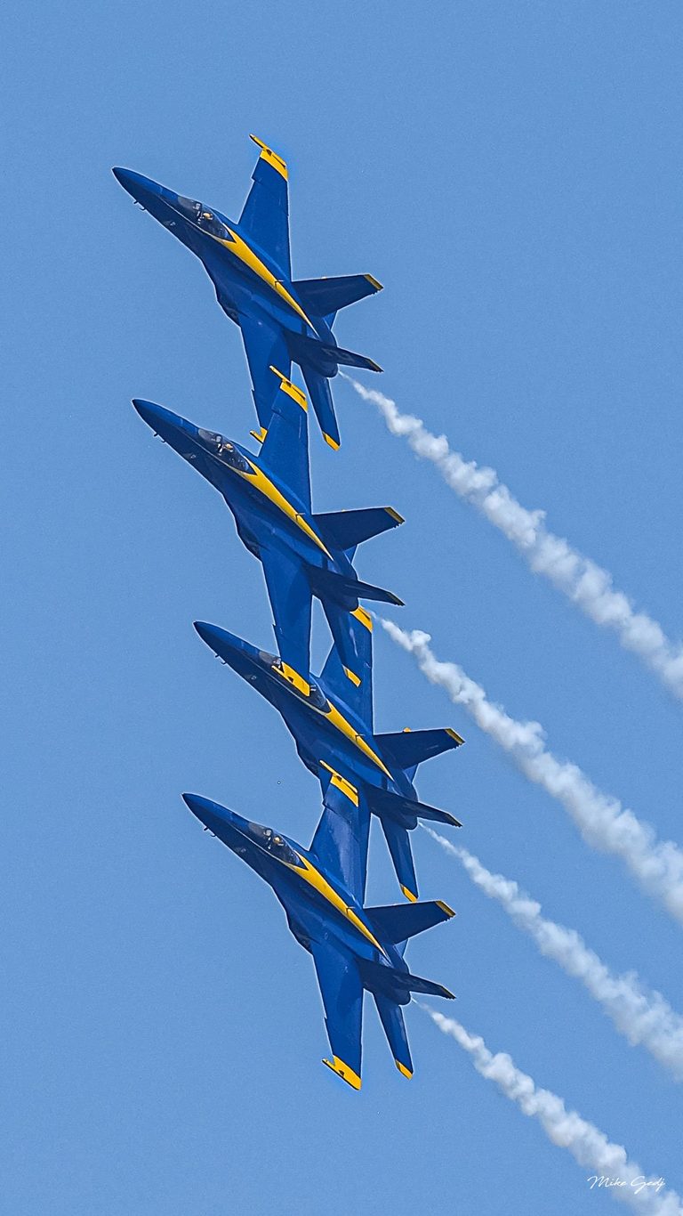Four blue and yellow jets bank in close formation against a clear blue sky, trailing white smoke toward the lower right.