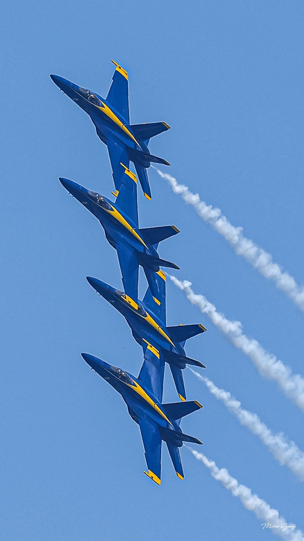 Four blue and yellow jets bank in close formation against a clear blue sky, trailing white smoke toward the lower right.