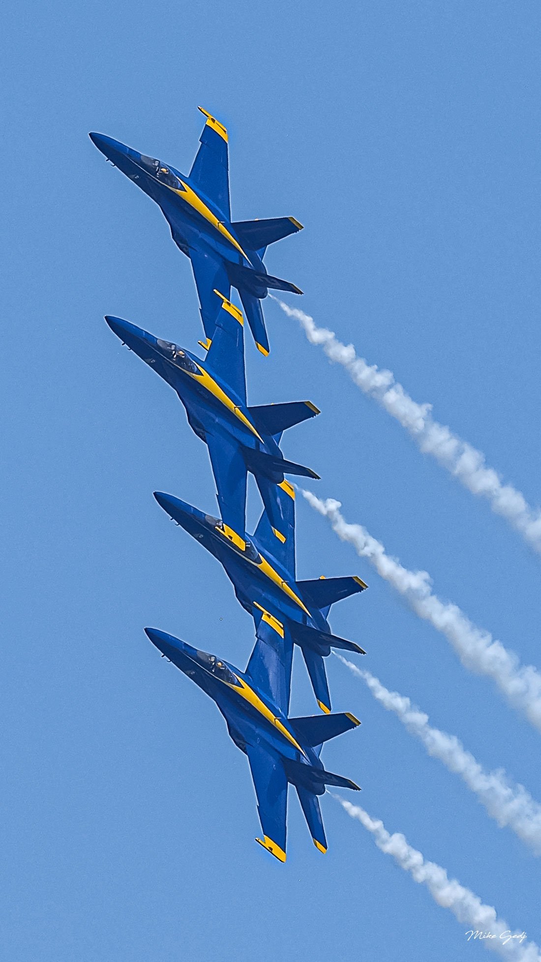 Four blue and yellow jets bank in close formation against a clear blue sky, trailing white smoke toward the lower right.