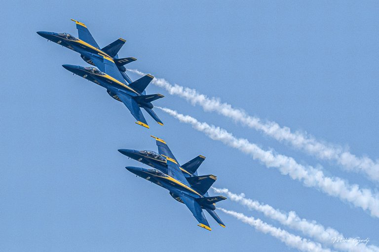 Four blue and yellow Blue Angels jets bank in formation against a clear blue sky, trailing white smoke toward the lower right.