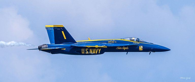 A blue and yellow U.S. Navy Blue Angels jet marked number 1 flies toward the right against a pale blue sky, trailing white smoke.