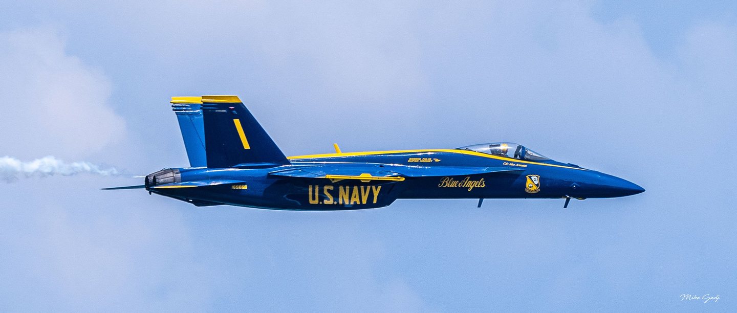 A blue and yellow U.S. Navy Blue Angels jet marked number 1 flies toward the right against a pale blue sky, trailing white smoke.