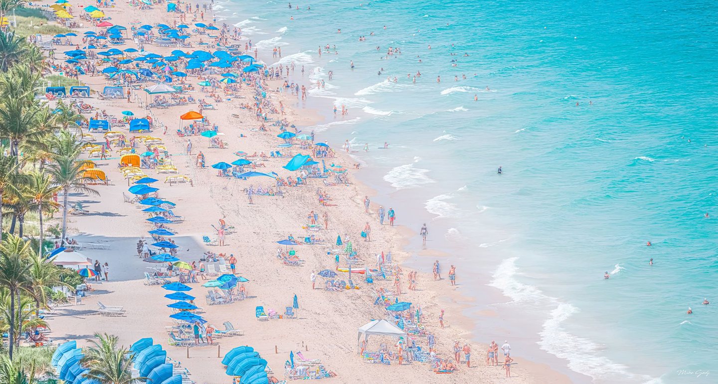 An elevated view shows a crowded sandy beach lined with palms, blue umbrellas and lounge chairs beside turquoise water dotted with swimmers.