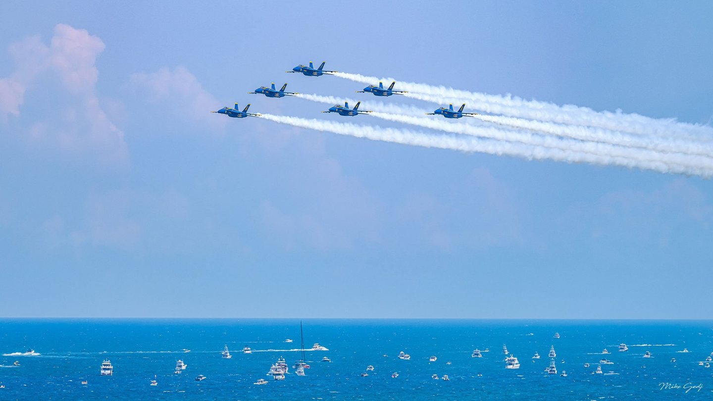 Six blue-and-yellow jets fly in formation toward the left, trailing white smoke across a pale blue sky above an ocean dotted with boats.