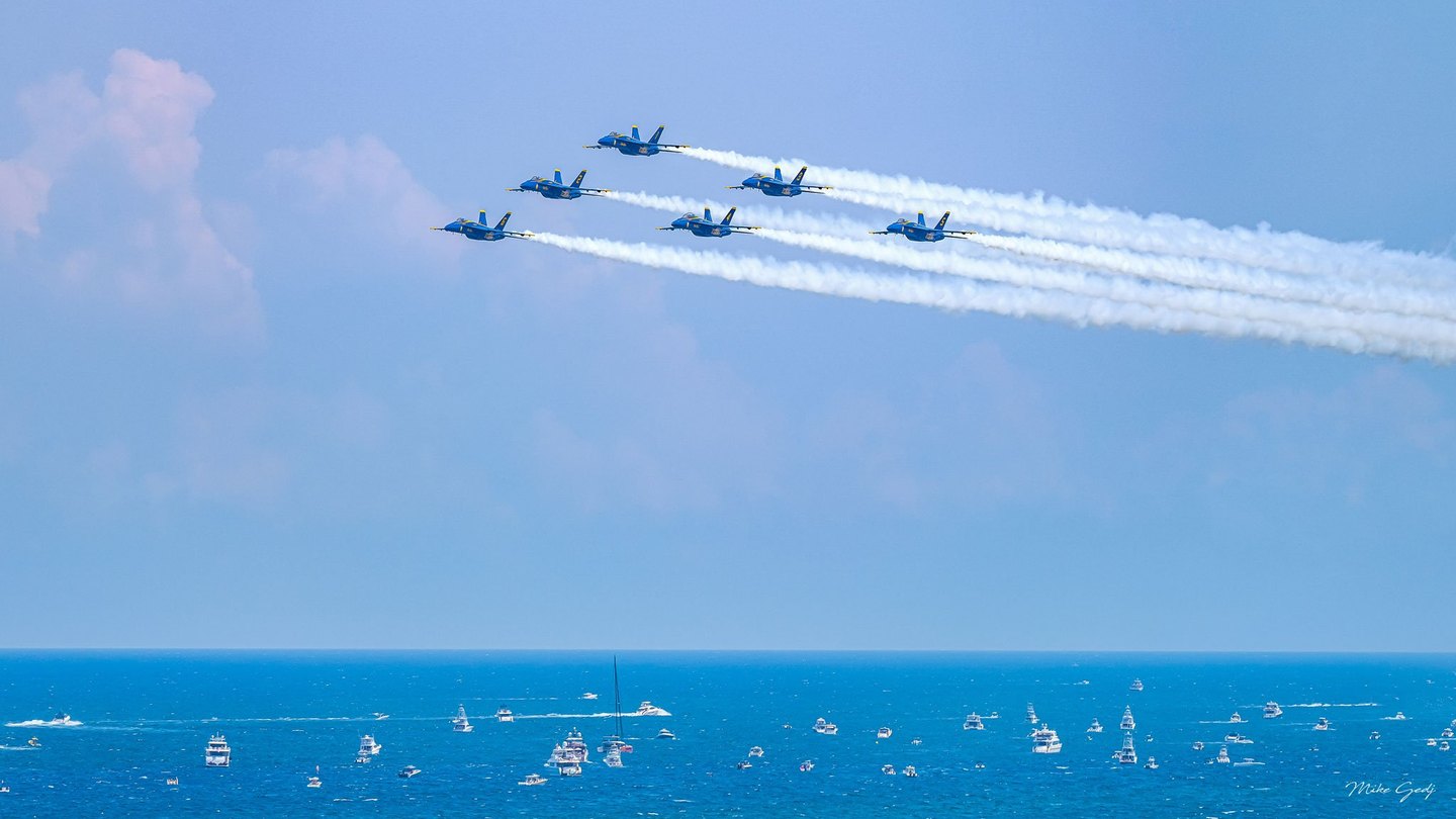Six blue-and-yellow jets fly in formation toward the left, trailing white smoke across a pale blue sky above an ocean dotted with boats.